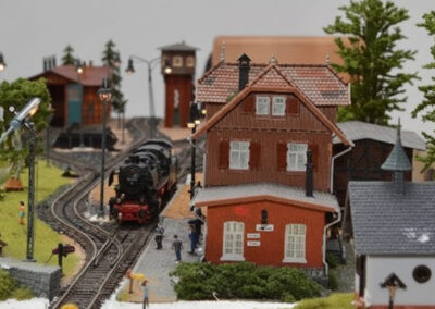 Detaillierte Modelleisenbahnanlage mit historischer Dampflokomotive auf geschwungenem Gleis, rotes Backsteinhaus mit Ziegeldach, Laternen und Miniatur-Bahnhofsszenerie