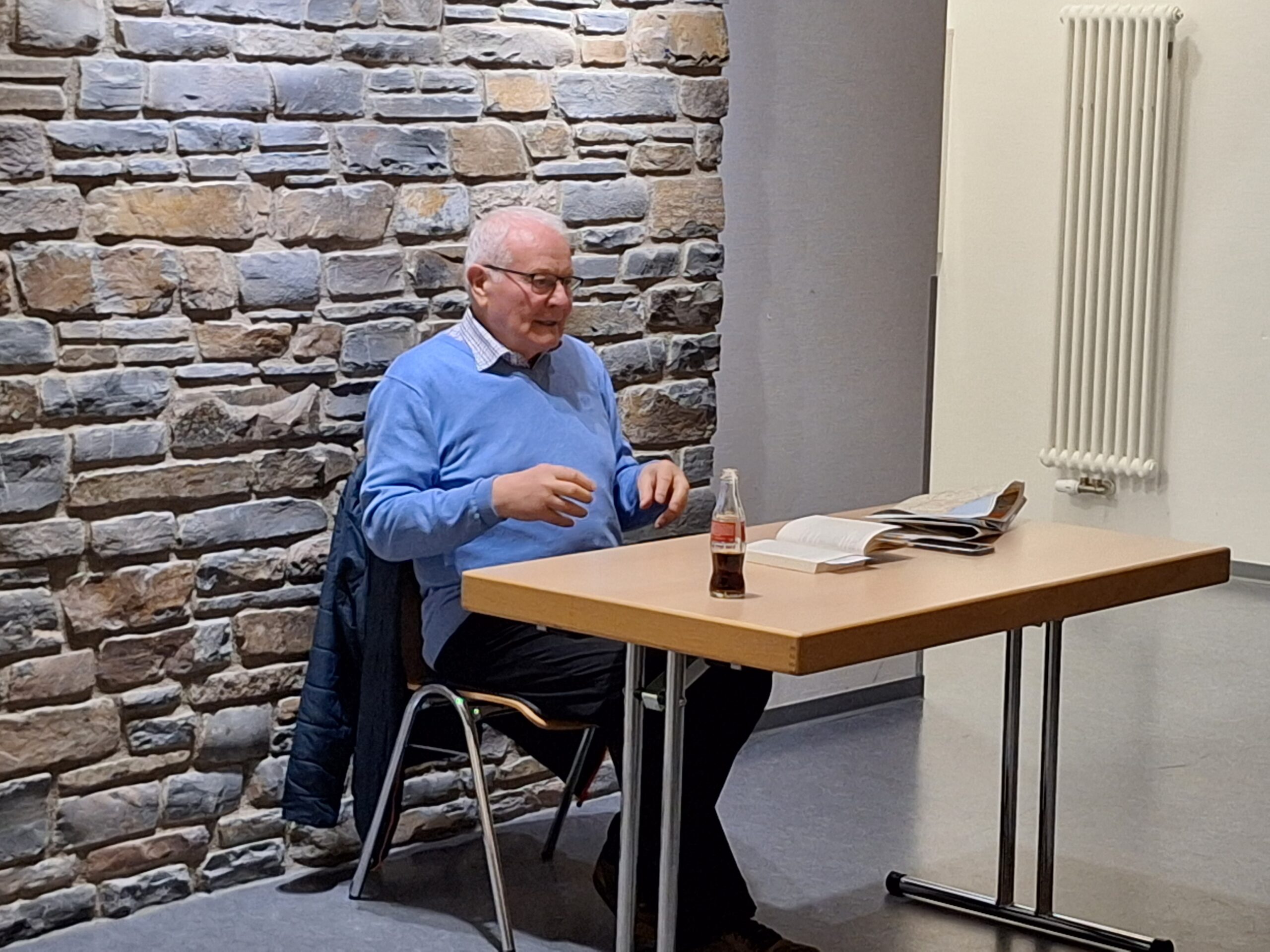 Werner Lutz am Tisch bei der Lesung, Älterer Mann in hellblauem Pullover hält Lesung an Tisch mit aufgeschlagenem Buch vor Natursteinwand