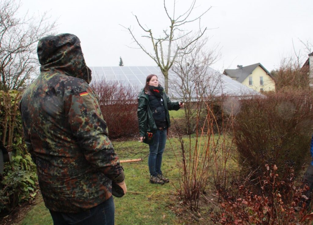 Zwei Personen im Garten bei winterlichem Wetter, eine in Tarnkleidung, die andere in grüner Jacke präsentierend, Solarpanels im Hintergrund